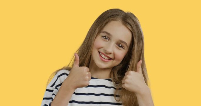 Close up of the blond pretty teen girl with long hair in the striped blouse giving her thumbs up and smiling while standing on the yellow wall background. Portrait