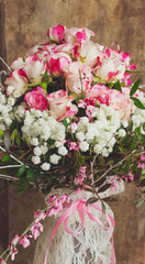 Close up view of a beautiful bouquet of pink roses and spring floral branches in a vase decorated with white lace and pink ribbon on wooden background, vintage style