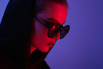 Portrait of fashion young girl in a black sweater with a hood and round sunglasses in red and blue neon light in the studio