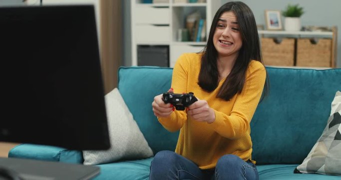 Caucasian young cheerful woman sitting at home on the couch and playing videogame with a joystick in front of the TV screen.