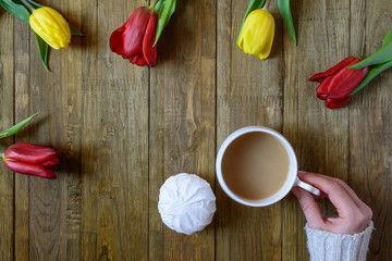 Flat lay of greeting card with flowers tulips and hand keeping white cup of coffee with milk on brown wooden background. Top view