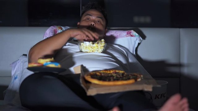 Young greedy overweight man eating popcorn and others junk foods likes pizza and donuts in dark bedroom while watching TV. Shot in 4k resolution