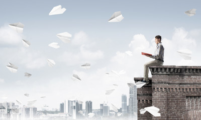 Handsome student guy on roof edge reading book and paper planes fly around