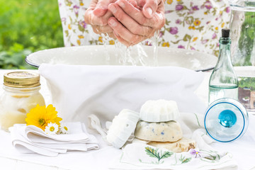Woman washes her hands in the white bowl outdoors, nearby are handmade soaps, flowers and cream, health and hygiene concept