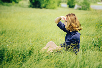 Portrait of a thoughtful girl in a meadow in the grass, sitting on a walk on a Sunny day covers his face with his hands from the bright light and looks into the distance.