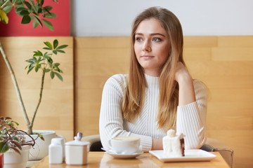 Obraz na płótnie Canvas Thoughtful beautiful woman focused pensively aside, drinks espresso and eats delicious cake in coffee shop, enjoys peaceful atmosphere, wears white jumper, has lunch alone. Free time concept
