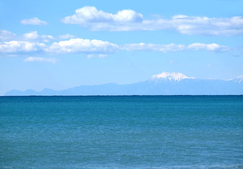 High mountains silhouettes with white snow cap on top far away on horizon in light haze after the sea under blue sky with white cumulus clouds