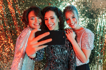 Beautiful young happy women friends posing over glitter wall background take a selfie by phone.