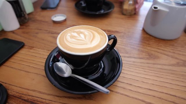 A smooth panning shot of a latte in a black cup & saucer. Latte art is featured on the face of the coffee. In the background are iPhones & tea facilities which are sat on a nice wooden table. 