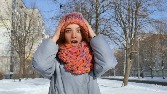 Young girl in warm coat and colorful knitted scarf putting on a hat standing outdoors during sunny day in spring on blue sky background