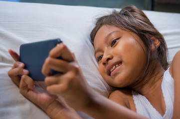 Obraz na płótnie Canvas happy and beautiful 7 years old child having fun playing internet game with mobile phone lying on bed cheerful and excited in young girl and technology concept