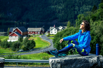 Portrait of young woman in blue clothes sitting on the stone and enjoying sunny day. Norway.