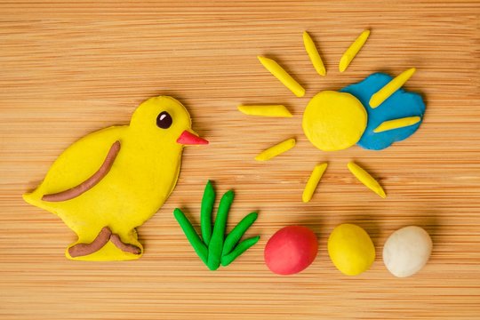 Funny spring Easter image made of plasticine, a yellow chicken, green grass, colorful eggs - red, yellow and white, golden sun shining over blue cloud on wooden background, warm colors