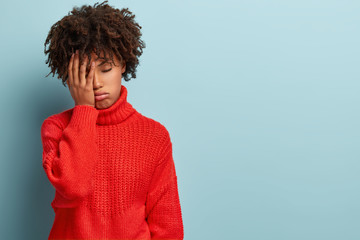 Discontent tired dark skinned woman covers face with hand, has tired expression, wears red sweater,...