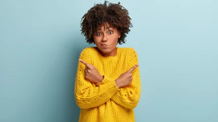 Poster Puzzled confused woman has hesitant expression, curly hairstyle, crosses hands over chest, points right and left in different sides, wears yellow sweater, being indifferent, models over blue wall © Wayhome Studio