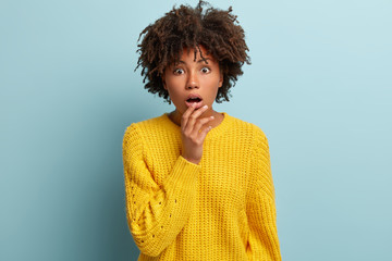 Frightened dark skinned young woman feels astonishment, keeps hand on dropped jaw, has widely opened eyes, dressed in yellow sweater, models over blue studio wall. People, shock and disbelief