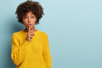 Studio shot of quiet dark skinned lady keeps fore finger over lips, makes silence gesture, gossips with friend, wears yellow sweater, says hush, isolated over blue studio wall with blank space - obrazy, fototapety, plakaty