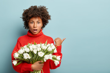 Shocked puzzled woman with Afro hairstyle, points away with thumb, carries beautiful white spring...