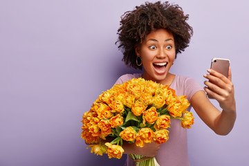 Shot of optimistic dark skinned lady holds cell phone for making selfie, has online translation in social networks, shares good impressions with followers which bouquet she recieved from colleagues