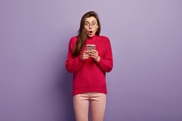 Half length shot of surprised woman holds mobile phone, writes notes in gadget, being distracted by bad news, has dark long hair, dressed in casual outfit, uses wireless internet. Modern communication