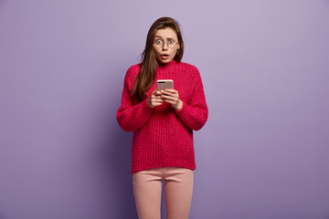 Upset puzzled woman uses mobile phone for online communication, types text messages on cellular, dressed in red sweater and trousers, isolated over purple background. Hmm, what strange notification