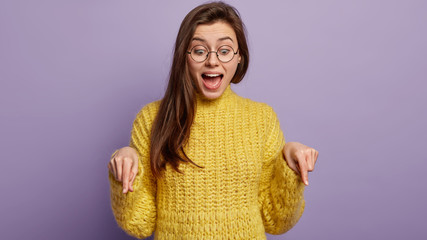 Photo of cheerful young European lady points down with both index fingers, smiles broadly as...