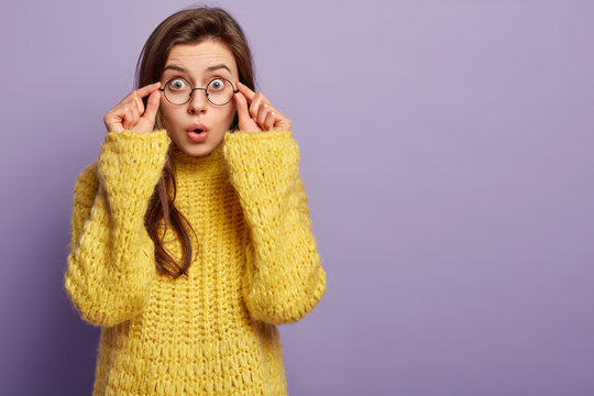 Emotive surprised student cann not believe she failed exam, touches rim of glasses, keeps lips rounded, wears yellow sweater, expresses shock and wonder, isolated over purple wall, free space