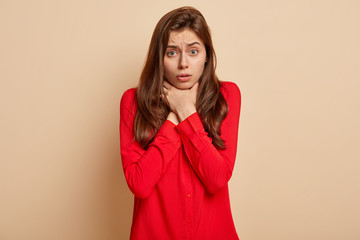 Frustrated young female keeps both hands on throat, suffers from asthmatic fit, can not breath well, wears red shirt, has problems with breathing, isolated over beige background. Cough attack