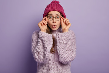 Headshot of scared speechless young woman touches rim of spectacles, keeps mouth widely opened wears fashionable winter clothes, isolated over purple background, sees incredible thing in front