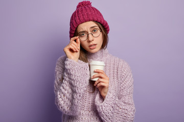 Portrait of unhappy Caucasian lady with sad tired expression, wears optical glasses, hat and knitted sweater, drinks coffee to go for feeling fresh and brave, dressed in winter outfit, poses indoor