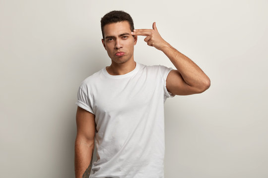 Horizontal shot of serious tired attractive guy shoots in temple with finger, demonstrates suicide gesture, wears white t shirt, fed up of long work, has strong body, isolated over white background.