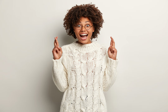 Positive emotional dark skinned young female crosses fingers for good fortune, has overjoyed facial expression, dressed in knitted white sweater, demonstrates strong desire for dreams come true