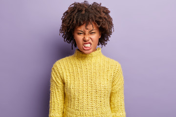 Furious outraged African American woman frowns face and clenches teeth, irritated because of much duties, has own style of clothing, isolated over purple background, annoyed with personal troubles.