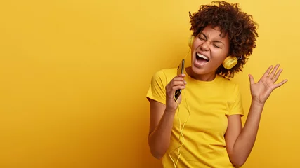 Deurstickers Yeah, I am pleased. Joyful lovely dark skinned woman raises hand in dance move, likes great sound in new headphones, sings loudly, carries cellular, smiles broadly, wears yellow casual clothes © Wayhome Studio