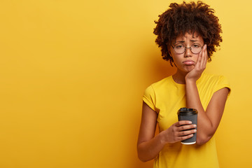 Upset dark skinned female feels sleepy and discontent to wake up early in morning, holds paper cup of coffee, wears transparent eyewear and yellow t shirt, keeps hand on cheek, being fatigue - obrazy, fototapety, plakaty