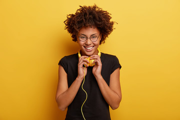 Isolated shot of optimistic woman with curly hairstyle, wears black t shirt and spectacles, feels...