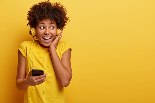 Indoor Shot Of Attractive Young Woman Listens Cool Song With Headphones And Mobile Phone, Has Nice Quality Of Sound, Focused Away, Has Broad Smile, Good Mood, Dressed In Casual Yellow Attire