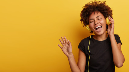 Emotional happy dark skinned lady enjoys every bit of pleasant melody, dances and gestures actively, wears stereo headphones, closes eyes, wears casual black t shirt, isolated on yellow wall - obrazy, fototapety, plakaty