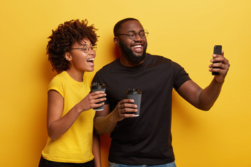 Indoor shot of happy ethnic girlfriend and boyfriend take selfie on modern gadget, laugh as being in good mood, drink coffee during spare time, express positive emotions, isolated on yellow wall