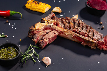 Set of grilled sliced T-bone steak with grilled corn, sauce, chilli, rosemary, salt, garlic on black background.