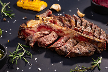 porterhouse t-bone steak is grilled sliced on a piece with grilled corn, sauce, chilli, rosemary, salt, garlic on black background. rustic style