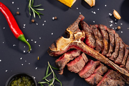 porterhouse t-bone steak is grilled sliced on a piece with grilled corn, sauce, chilli, rosemary, salt, garlic on black background. rustic style
