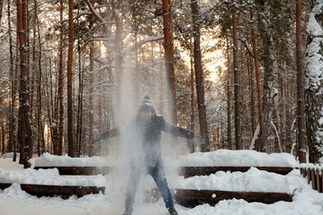 A young man in a winter coniferous forest is standing and catching with his hands the snow falling from the branches of trees looks like he sparkles in the sun and smiles.