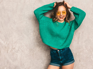 Beautiful sexy smiling gorgeous girl in green trendy sweater. Woman posing near gray wall in round sunglasses. Model having fun and showing her tongue