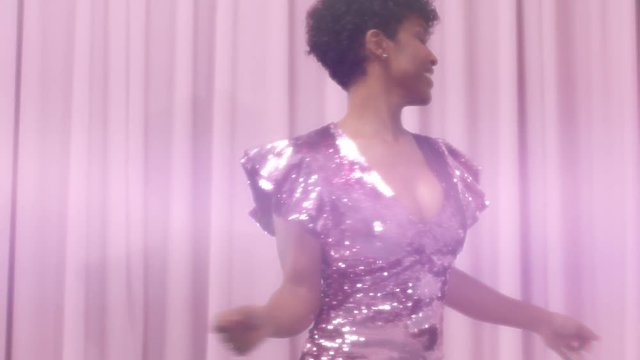 black mixed race woman with short haircut and curly natural hair wears sequin sparkly dress in pink. Dancing womn and sparkly sequin dress makes lens flair