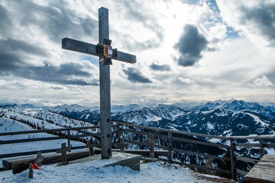 A picture from the sunny day in austrian Alps. The visibility is great, the nature is very nice. The peaks of the mountains are hiding in some clouds. A wooden cross memorial is in front. 