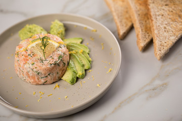 Rohes Lachstatar mit Lachs, Dill, Zitrone, Avocado und Vollkorn Toast Brot auf Porzellan Teller und Marmor Hintergrund