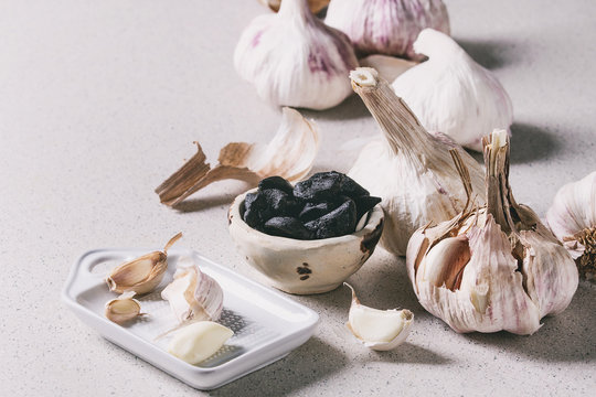 Variety of fresh organic garlic bulbs whole and peeled and cloves of black fermented garlic with ceramic grater over grey spotted background. Close up