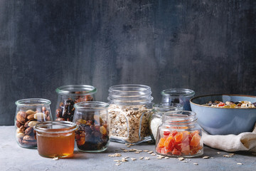 Variety of dried fruits, nuts, honey and oat flakes in bowl and different glass jars for cooking homemade healthy breakfast muesli or granola energy bars on grey blue table. - Powered by Adobe