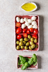 Mozzarella, cherry tomatoes, olives antipasto appetizers served in pink ceramic rectangular plates with olive oil, basil in row over grey texture background. Flat lay, space